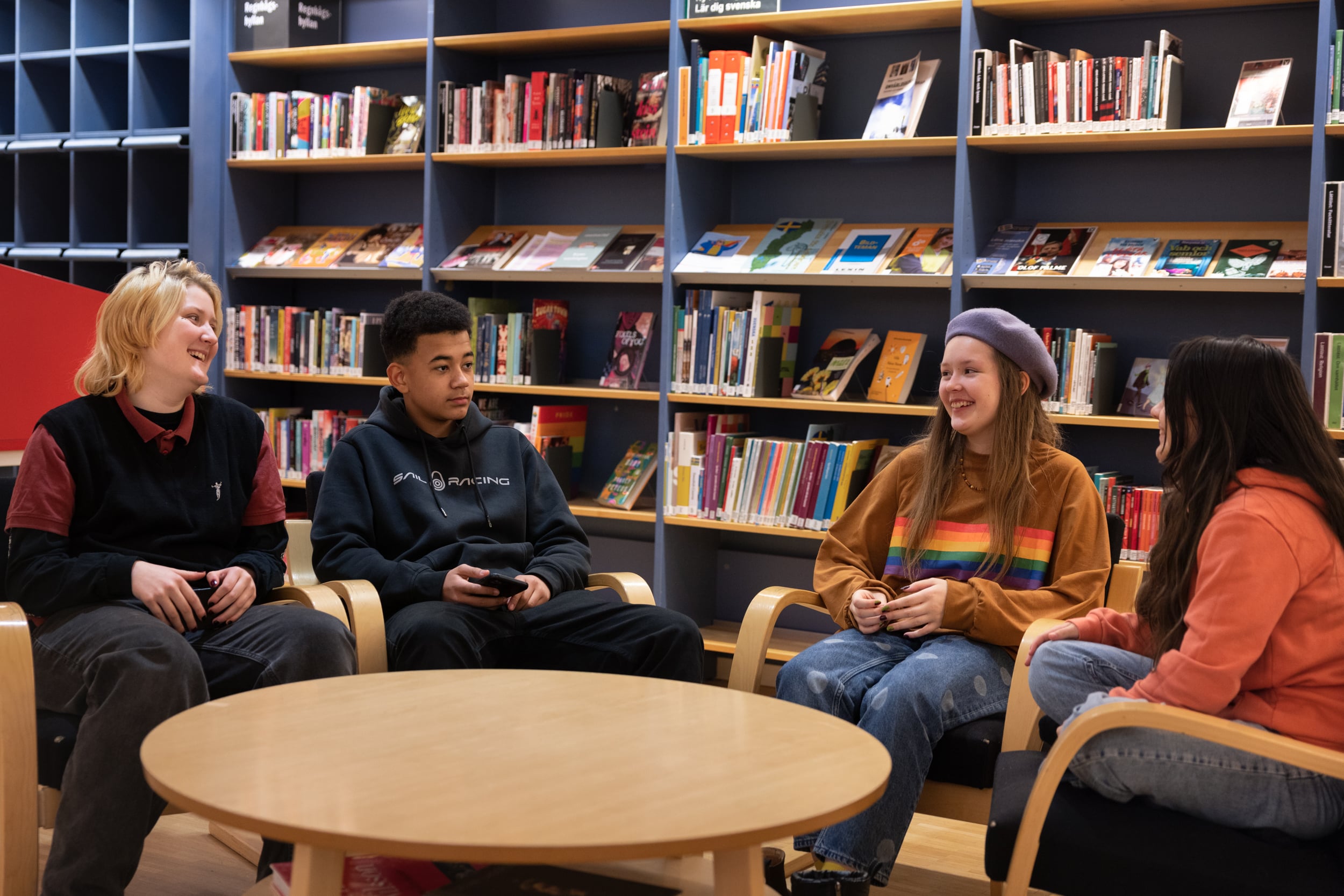 Fyra ungdomar sitter vid ett bord i ett bibliotek. De har roligt och pratar.