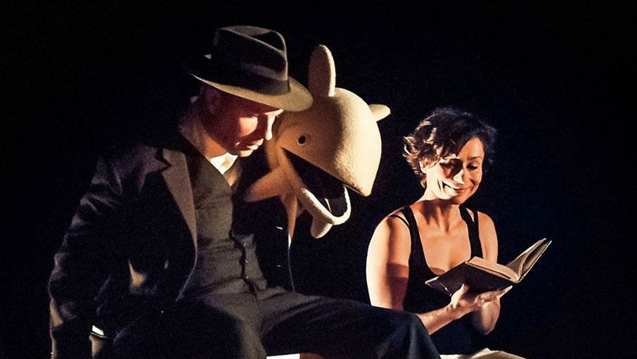 En man i kostym och hatt sitter bredvid en kvinna som läser i en bok på en teaterscen
