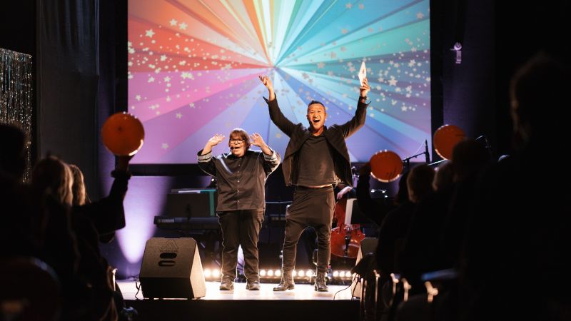 Scenbild från Funkisfestivalen där två personer står på scenen med armarna uppsträckta inför en publik. I bakgrunden syns färgglada ljuseffekter och stjärnor.
