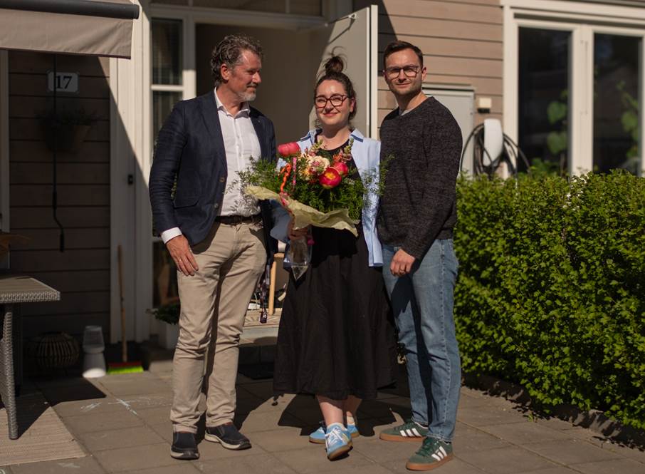 Den 22 maj uppvaktades Katie, Adam och deras son Noah av kommunstyrelsen ordförande Sven Gustafsson (M) Bild på tre personer utanför ett hus. Kvinnan på bilden har en stor sommarbukett i famnen.