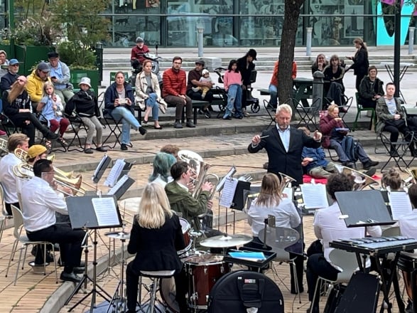 Människor sitter på ett torg och lyssnar på en orkester som spelar.
