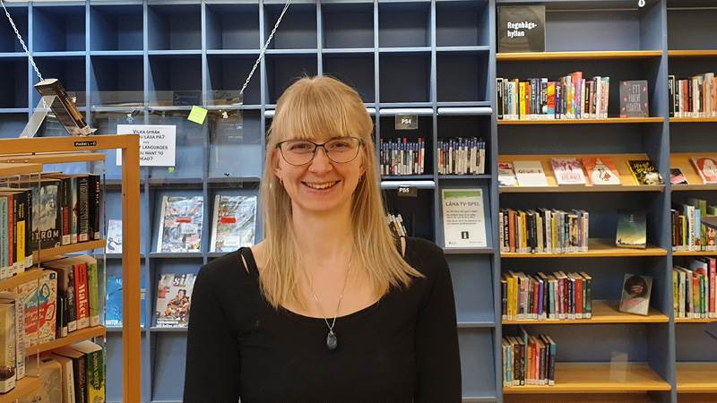 Janna Berg är samordnare för Regnbågsveckan i Haninge. Här står hon framför regnbågshyllan på biblioteket i Brandberget. Janna Berg står framför bokhyllor och ler mot kameran