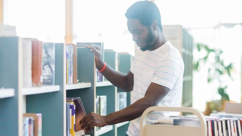 En ung man står vid en bokhylla och ställer in böcker från en vagn