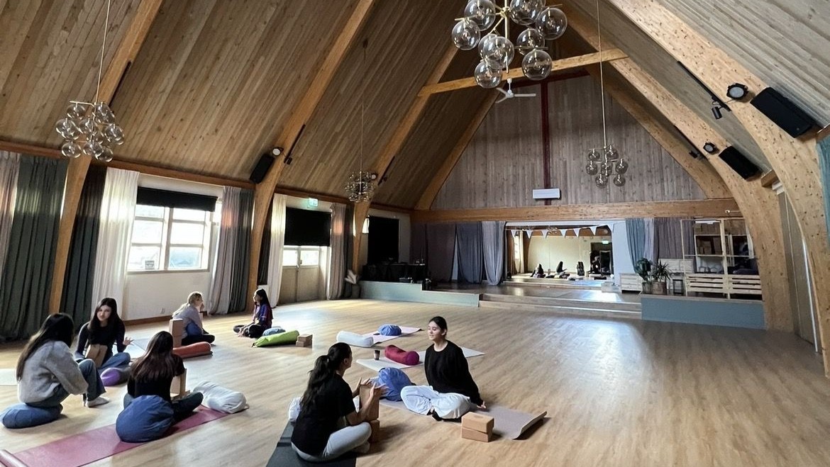 Personer sitter på yogamattor i en stor sal med högt trätak och stora fönster. Kuddar och yogablock ligger utplacerade på golvet.