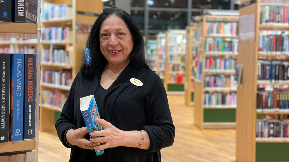 Kvinna står i ett bibliotek. Hon håller i en bok och ler.