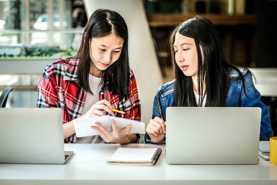 Två flickor i högstadieåldern sitter tillsammans med varsin dator vid ett bord och studerar.