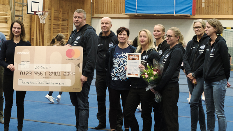 Två personer håller i en stor prischeck framför en grupp med människor i en idrottshall.