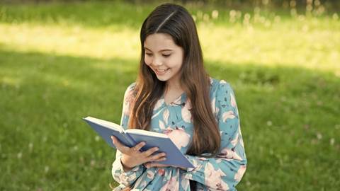 En flicka tittar glatt ner i sin bok, på en grön gräsmatta