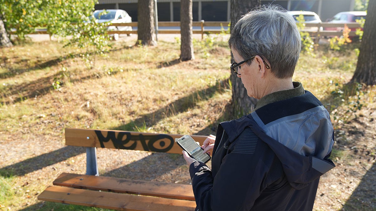 Kvinna står vid en nedklottrad parkbänk med en mobiltelefon i handen