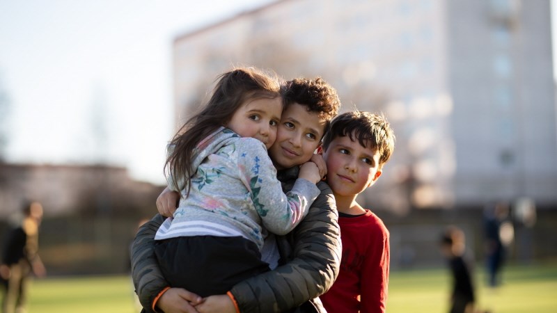 Tre barn håller om varandra på en skolplan, en flicka till vänster, en pojke i mitten och en pojke längst till höger.