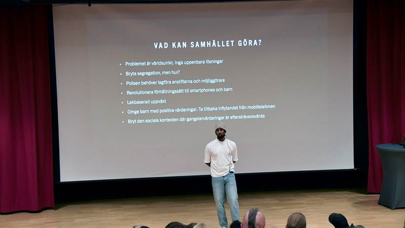 Föreläsaren Luay Mohageb håller en presentation på scen framför en stor skärm med text om förslag på samhällsåtgärder.