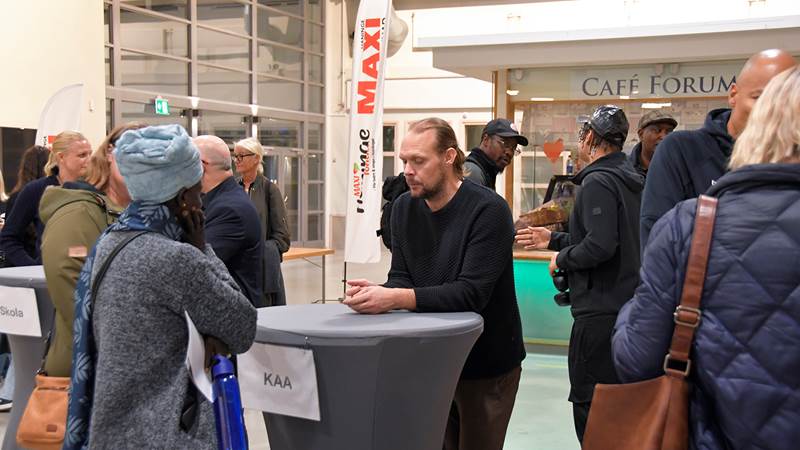 Deltagare samtalar vid höga bord. En skylt med texten 'KAA' och en banner från Maxi Stormarknad Haninge syns i bakgrunden, som sponsor av fikat från kvällen.