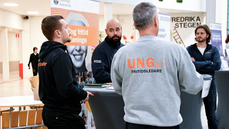 Grupp med ungdomsledare i tröjor märkta 'UNGish FRITIDSLEDARE' står vid informationsbord.