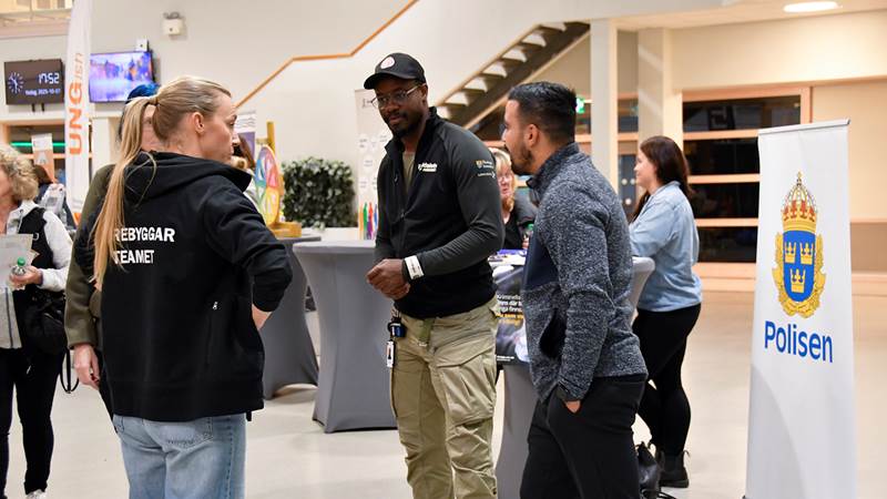 Tre personer samtalar vid ett evenemang, varav en bär jacka med texten "Förebyggarteamet" och en banner från Polisen syns i bakgrunden.