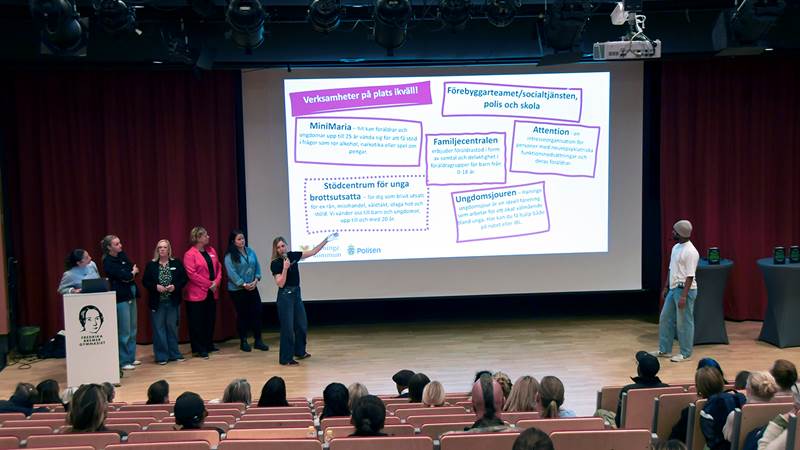 Presentation på scen med flera personer och en skärm som visar vilka lokala stödverksamheter som är på plats, inför en sittande publik.