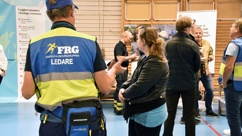 En person i väst märkt 'FRG FRIVILLIGA RESURSGRUPPEN LEDARE' samtalar med deltagare vid invigningen av trygghetspunkter.