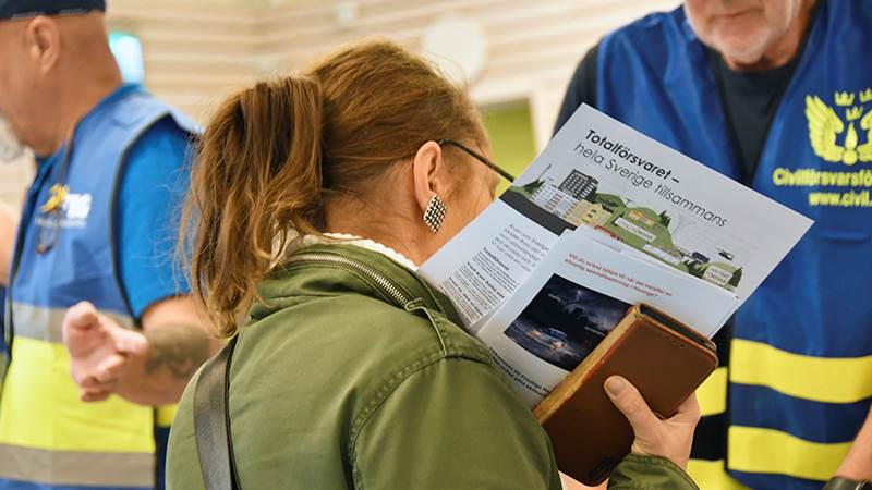 En person läser en broschyr med titeln 'Totalförsvaret – hela Sverige tillsammans', medan två personer i blå västar med civilförsvarstext syns i bakgrunden.
