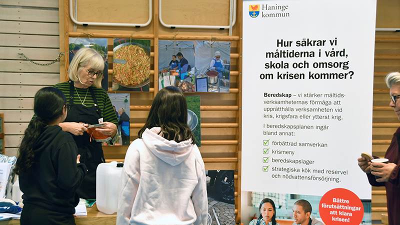 En kvinna, kommunens koststrateg, samtalar med två yngre personer framför en affisch från Haninge kommun om hur måltider säkras inom vård, skola och omsorg vid kris.