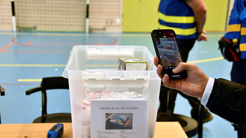 En plastlåda med etiketten 'Vad bör du ha i din krislåda?' visas upp. En person fotograferar innehållet med sin mobiltelefon.