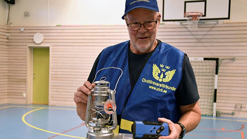En representant från Civilförsvarsförbundet håller upp en oljelampa och en annan krispryl, iförd blå väst med texten 'www.civil.se'.