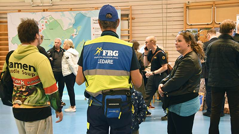 Personer samlade vid ett informationsbord under invigningen av trygghetspunkter. En person bär väst med texten 'FRG LEDARE' och en stor karta med trygghetspunkterna syns i bakgrunden.