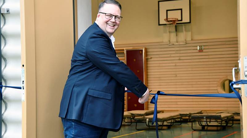 Jens Davidson (KD), kommunalråd i Haninge kommun, klipper ett blått band vid invigningen av trygghetspunkterna. I bakgrunden syns en gymnastiksal med uppställda fältsängar och en basketkorg.