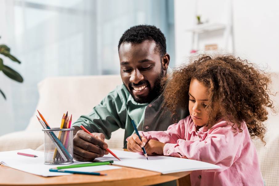En pappa med sin dotter sitter vid ett bord och ritar med färgpennor.