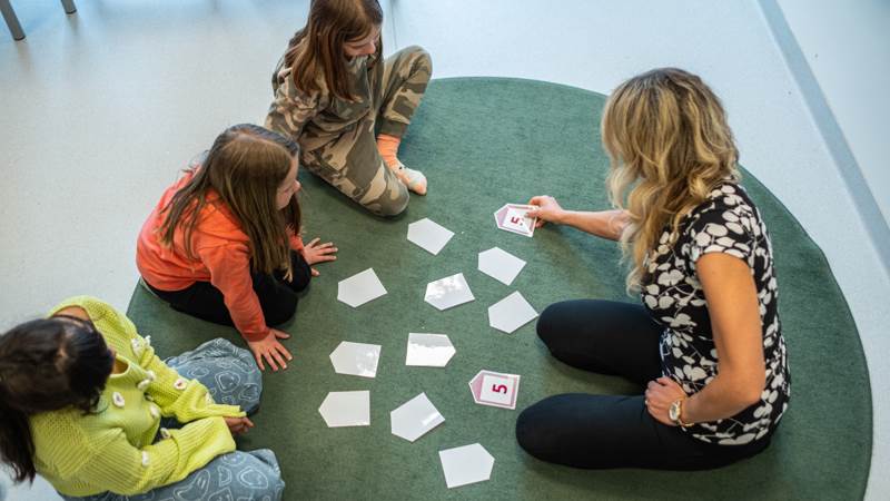Lektion i Vikingaskolans sverigefinska klasser. Tre barn och en lärare sitter på en grön matta och tittar på kort som ligger utspridda på mattan.