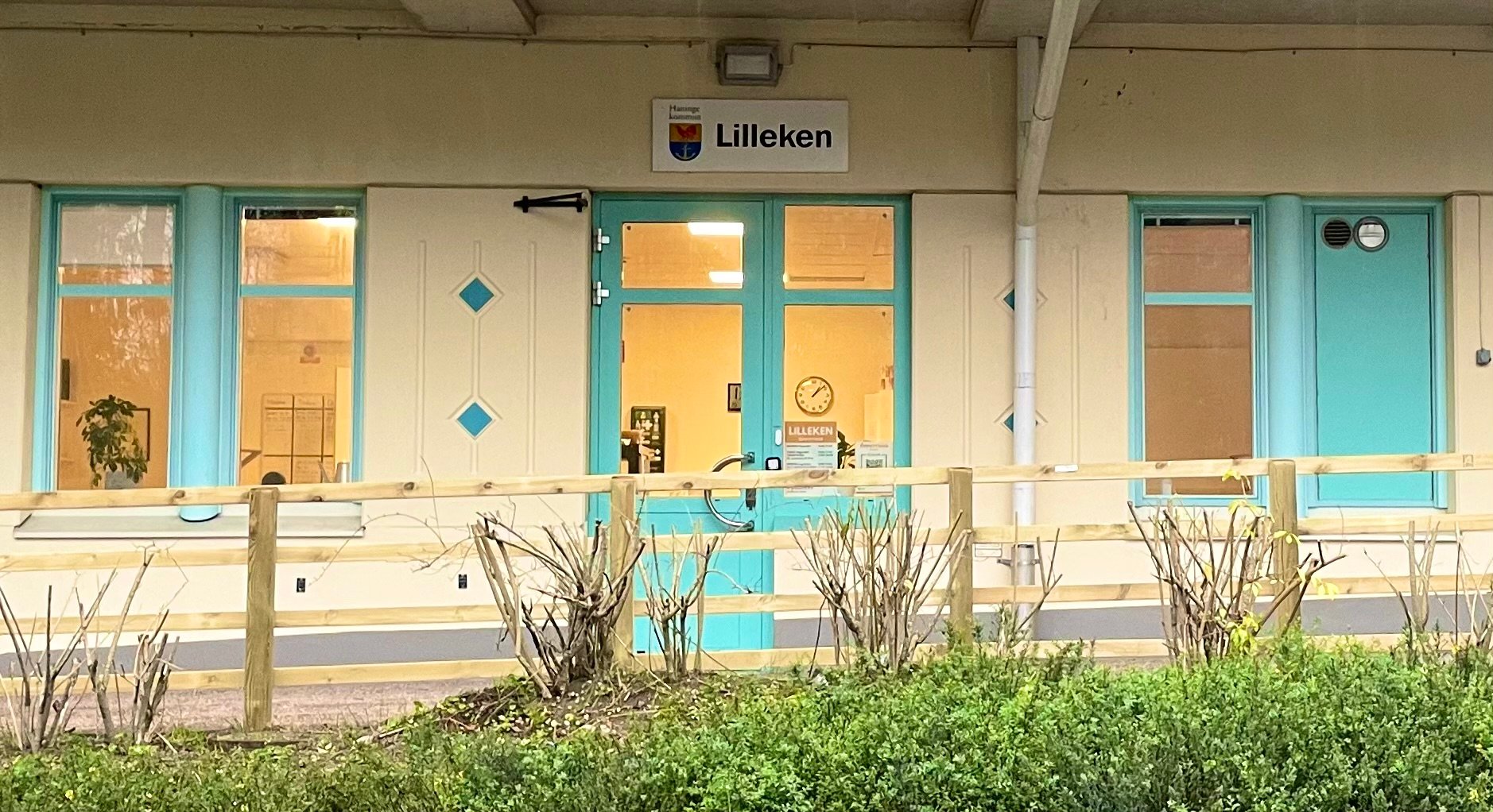 Entré till förskolan Lilleken i Haninge kommun. Byggnaden har ljus fasad med turkosa dörrar och fönsterkarmar. Ovanför dörren sitter en skylt med Haninge kommuns logotyp och texten ”Lilleken”. Framför entrén finns ett trästaket och några buskar.
