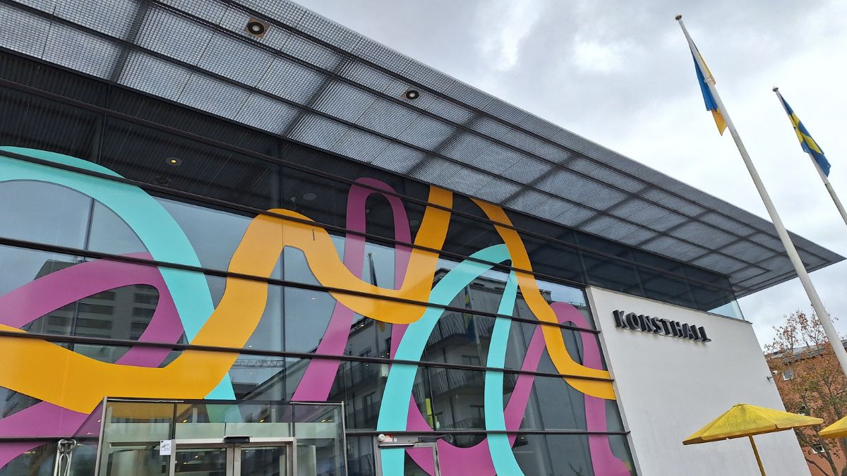 Exteriörbild av Haninge kulturhus med Konsthallens entré. Byggnadens glasfasad pryds av färgstarka mönster och svenska flaggor syns utanför.