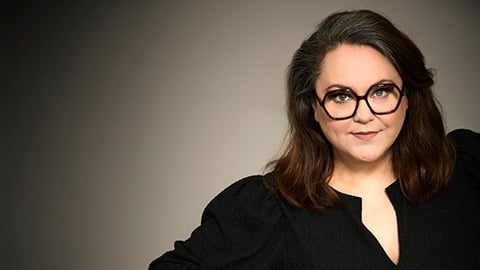 Porträttfoto av en person med glasögon och mörkt hår mot en ljusgrå bakgrund.