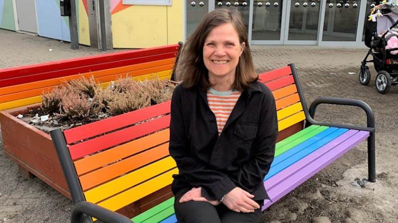 Henrika Åström sitter på en av de regnbågsfärgade bänkarna på Hurtigs torg. En kvinna sitter på en regnbågsfärgad bänk och ler mot kameran