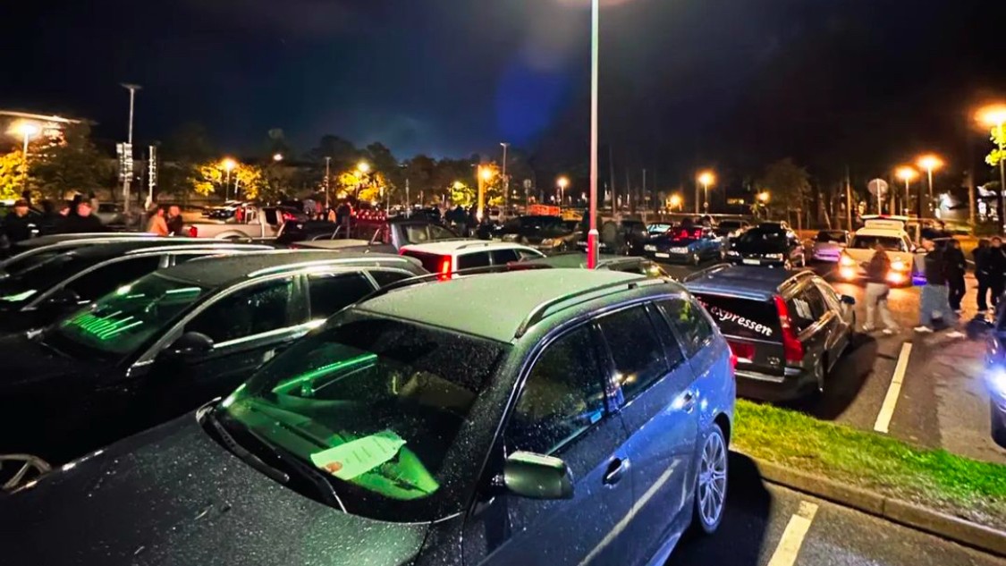 Flera epa‑traktorer står parkerade på en upplyst parkering under kvällen, med grupper av ungdomar som pratar och umgås i bakgrunden.