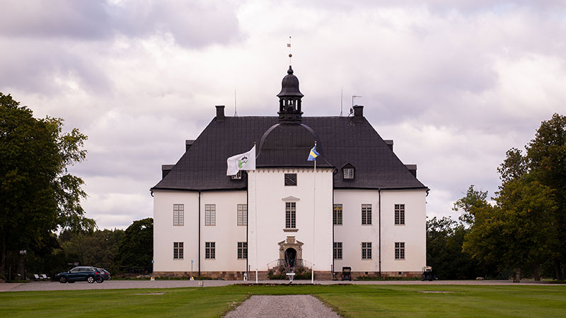 Årsta slott sett framifrån. Den ljusa slottsbyggnaden med mörkt tak ligger i en parkmiljö med gräsmattor, träd och en grusgång som leder fram till entrén.