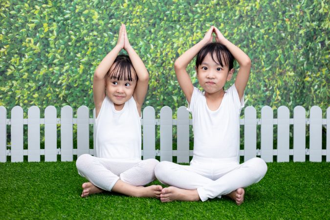 Två barn sitter på gräs och gör en yogaposition med armarna ovanför huvudet