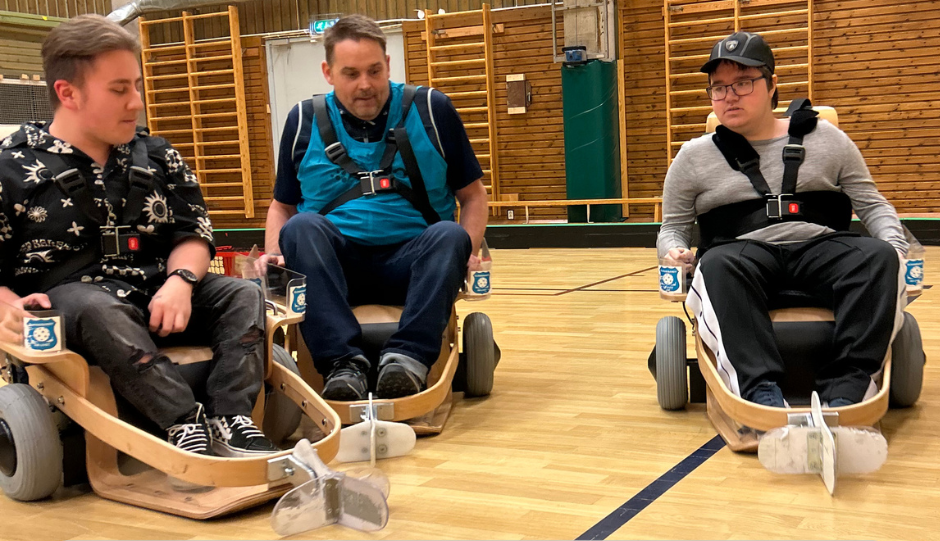 Tre personer sitter i varsin elrullstol som styrs med joystick i en idrottshall.