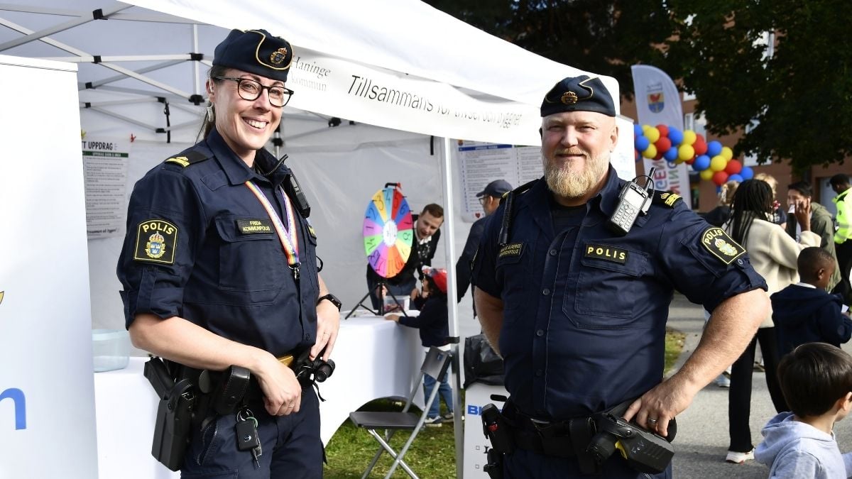 Två polisanställda i uniform står framför ett informationstält vid ett utomhusevenemang. I bakgrunden syns besökare, ett lyckohjul och färgglada ballonger.