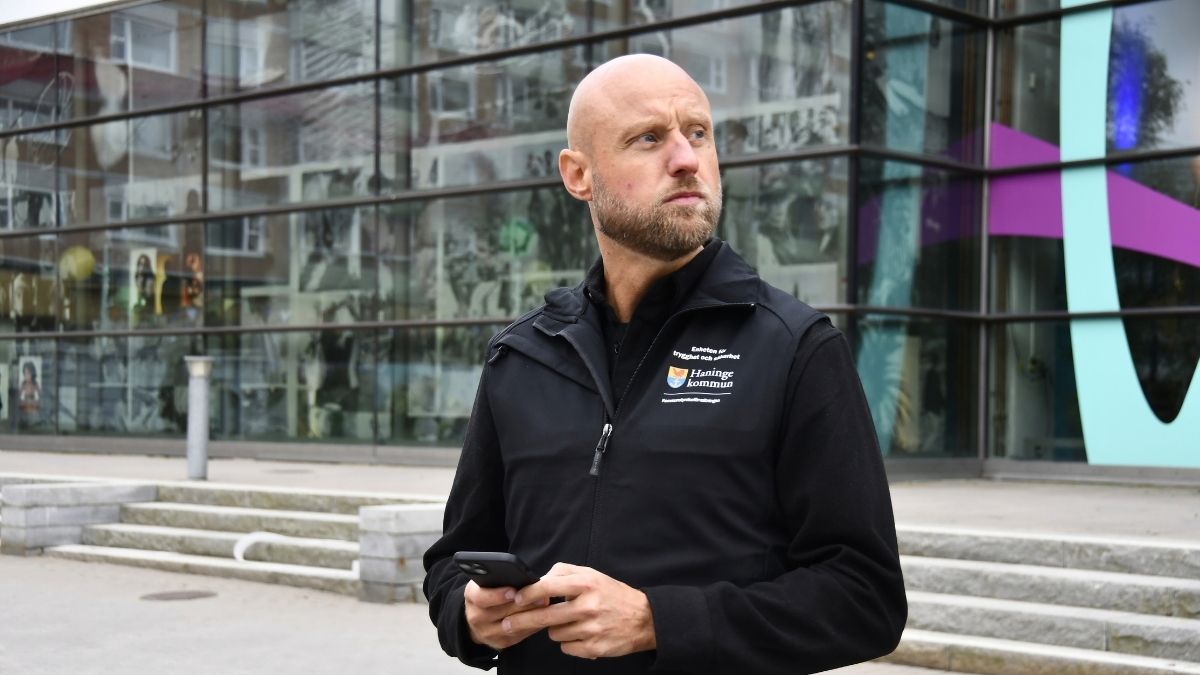 Person står utomhus framför en modern glasfasad och håller en mobiltelefon i händerna. Personen bär en svart väst med Haninge kommuns logotyp och text om trygghet och säkerhet.