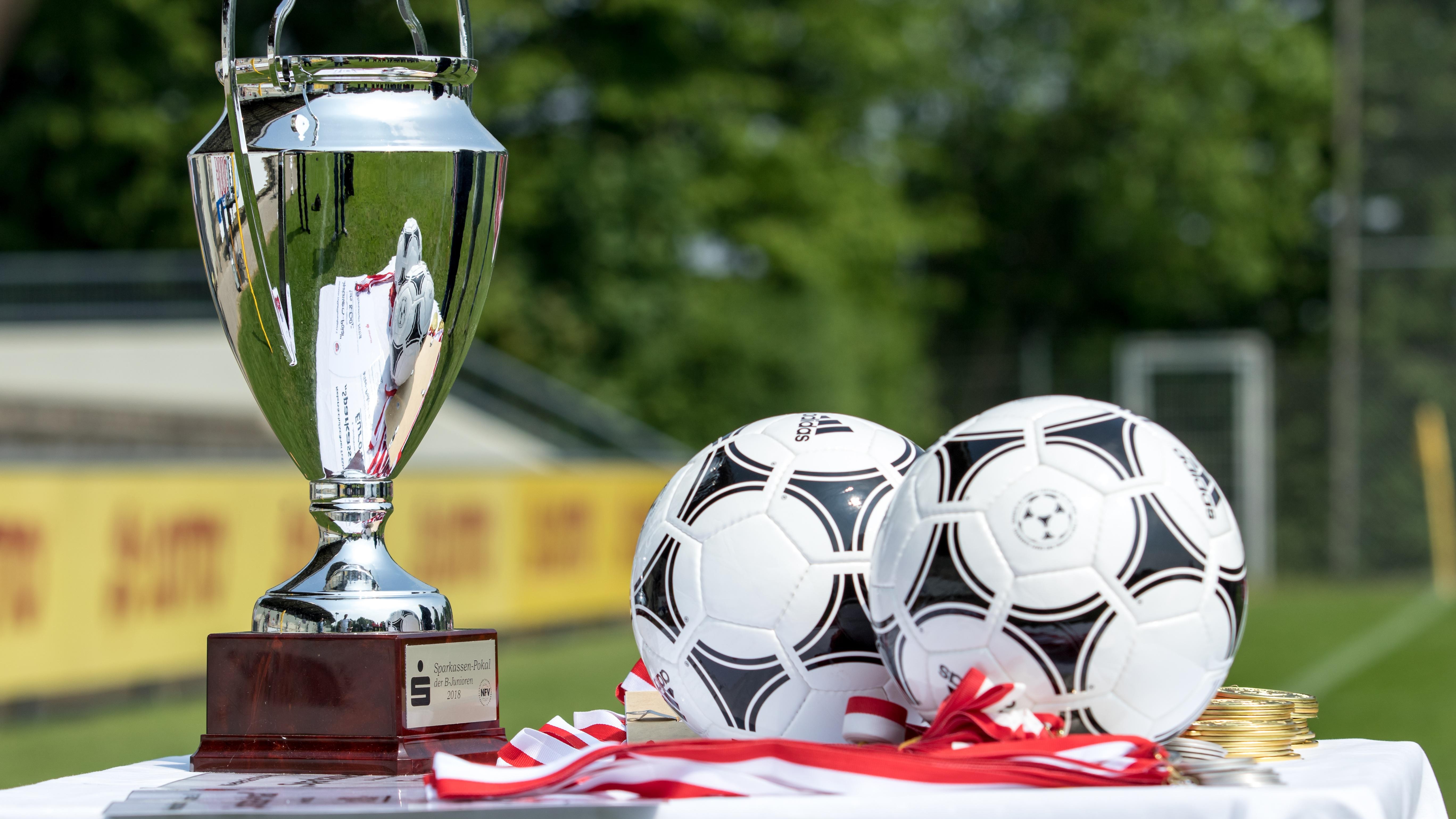 En pokal, två fotbollar och medaljer ligger uppställda på ett bord vid en fotbollsplan.
