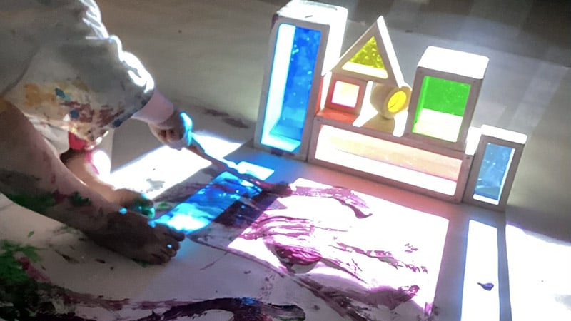 Solljus skiner genom färgade prismor. Ett barn sitter på golvet och målar.