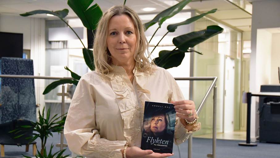 Författaren håller upp sin bok med båda händerna. Boken har en mörk omslagsdesign med titeln "Flykten till verkligheten". Kvinnan bär en ljus, långärmad blus med volanger vid bröstet och ärmarna, samt ett armband med pärlor. Bakgrunden är inomhusmiljö med metallräcken och suddiga detaljer.