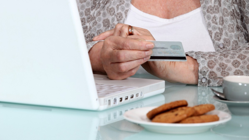 En äldre person sitter vid ett bord med en vit bärbar dator och håller ett bankkort i handen, redo att göra en betalning online. På bordet står en vit kopp med fat och en tallrik med kakor.