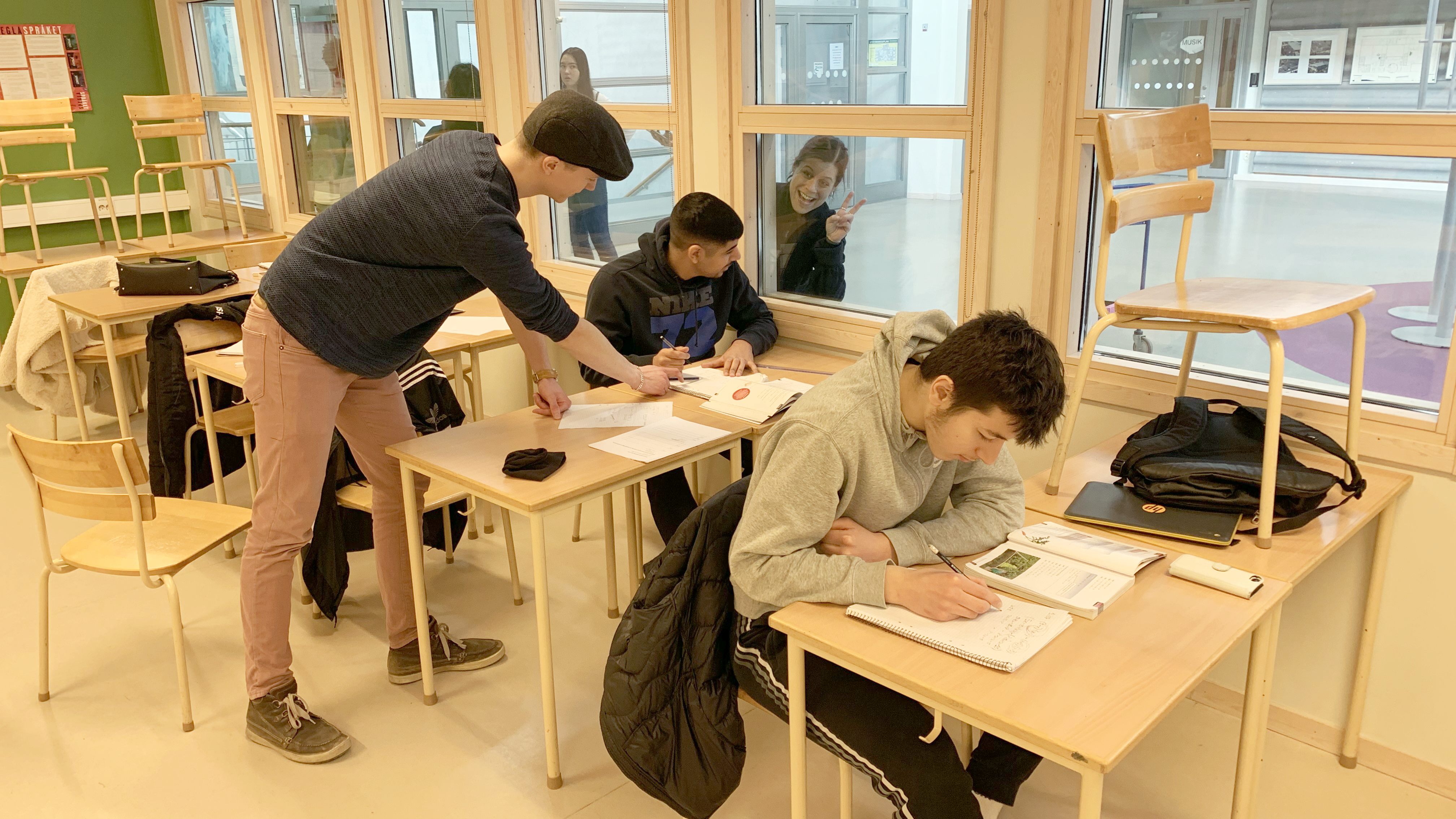 En lärarstudent står lutad över eleven och hans uppgifter medan en annan lärarstudent gör segertecken bredvid skolbänken, på andra sidan glaset. Framför sitter ytterligare en koncentrerad elev med sina uppgifter.
