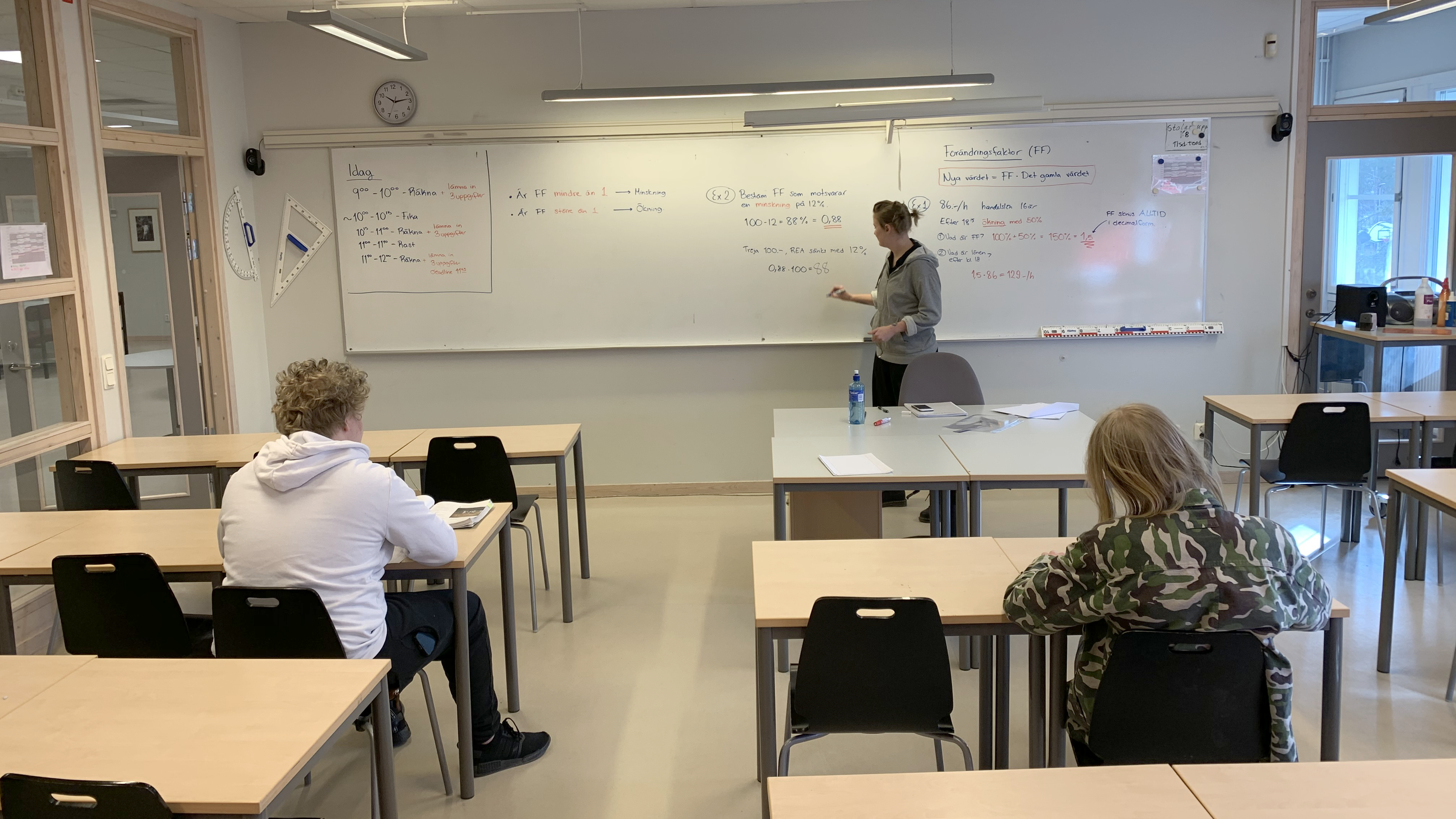En lärarstudent står vi tavlan i ett klassrum och förklarar en uppgift för två elever som sitter vid sina skrivbord.