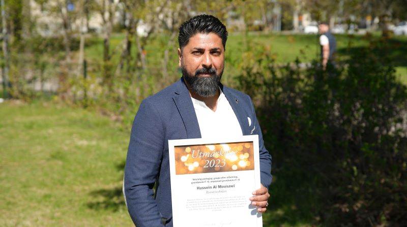 Hussein Al Mousawi, socialpedagog på Runstensskolan. Porträttbild på man i blå kostym hållande en utmärkelse. Stå utomhus i solen.