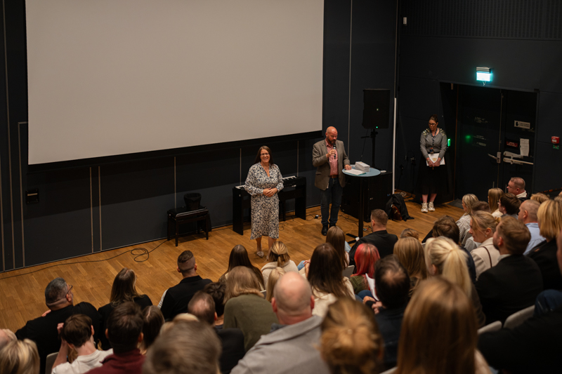 Flera personer sitter i publiken och en kvinna och en man står på scenen. 
