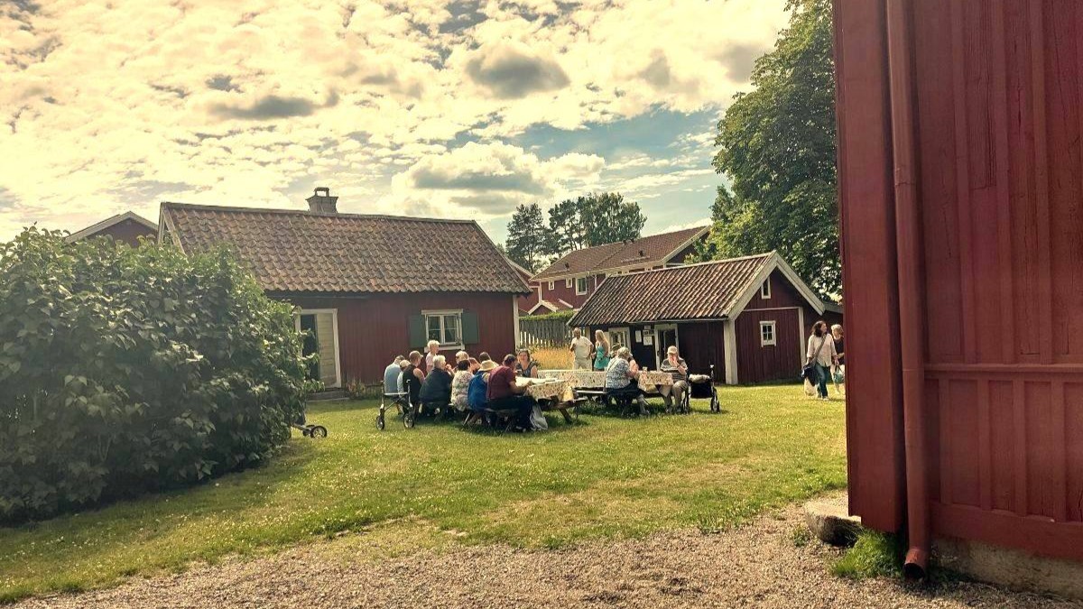 Människor sitter och fikar vid sommarcafét vid Tinghuset, omgivet av röda hus med vita knutar i en grönskande miljö.