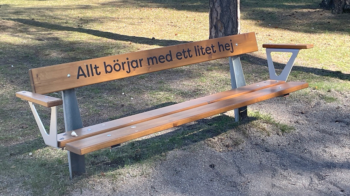 Träbänk i en park. På en av brädorna står "Allt börjar med ett litet hej"