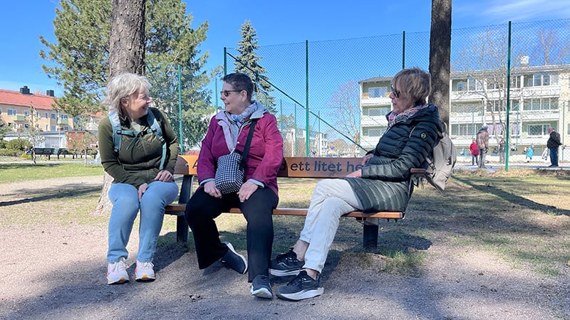 Tre glada kvinnor sitter på en parkbänk och pratar med varandra.