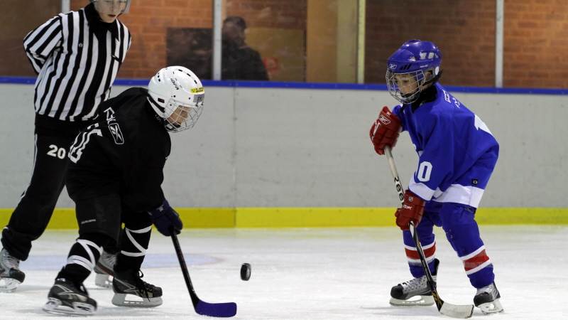 Två barn i hockeyutrustning spelar hockey medan domaren ser på de spelande.